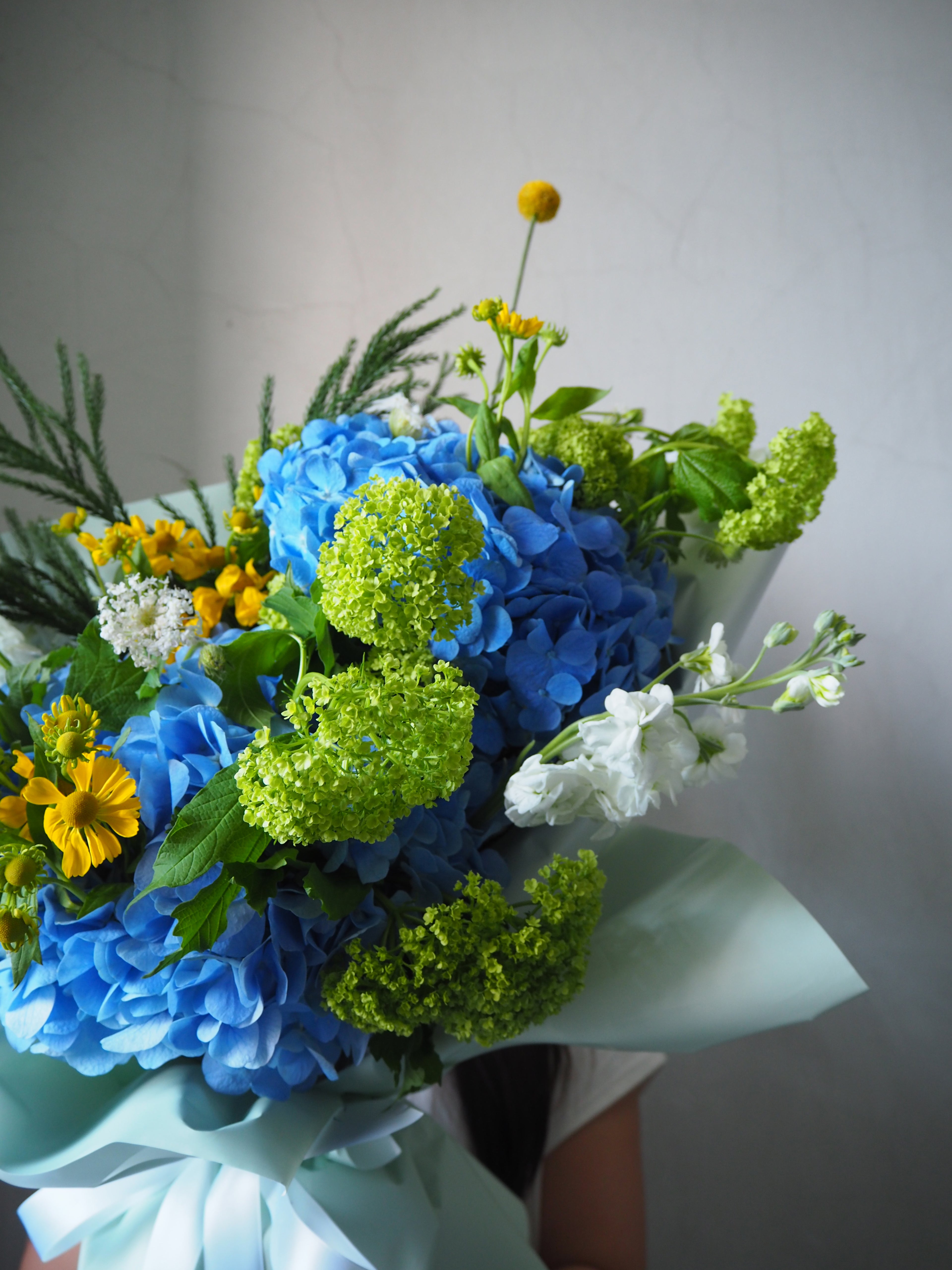 Bouquet of blue hydrangeas, green astilbes, and yellow daisies against a neutral background