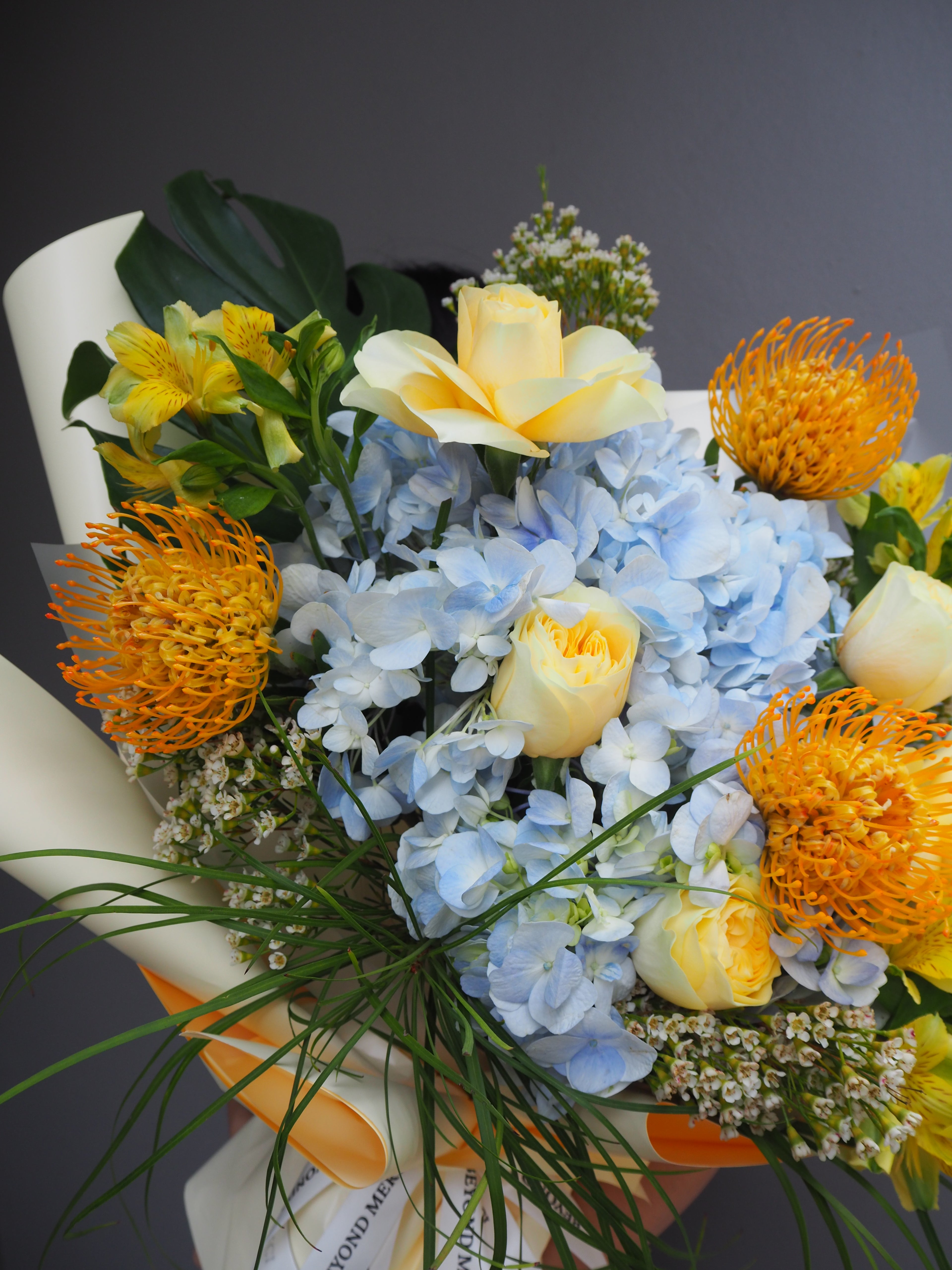 A luxe bouquet of flowers with blue hydrangeas, yellow roses, and yellow proteas on a dark background.