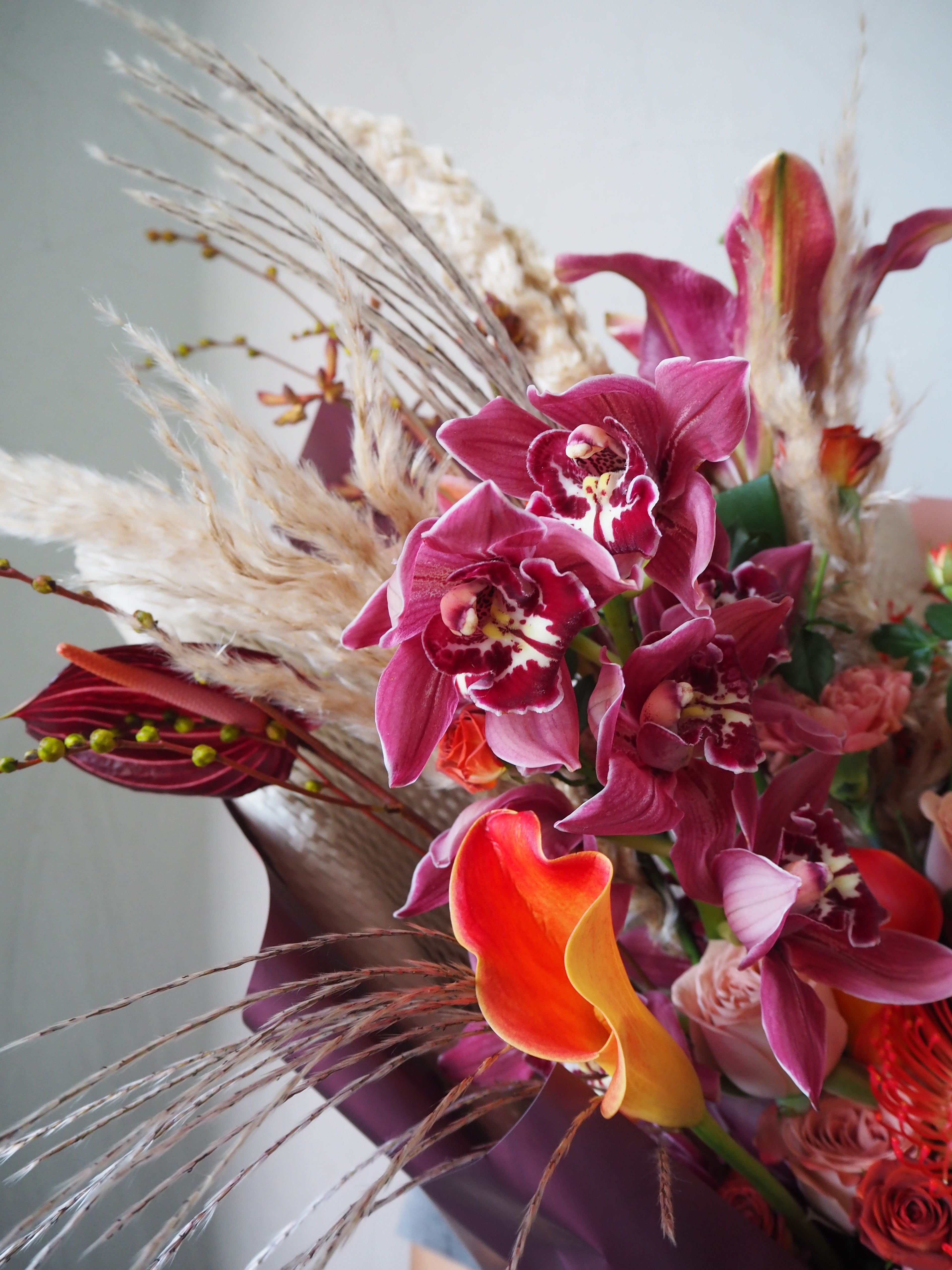 Close-up of a vibrant bouquet with pink orchids, red flowers, and dried elements on a light gray background.