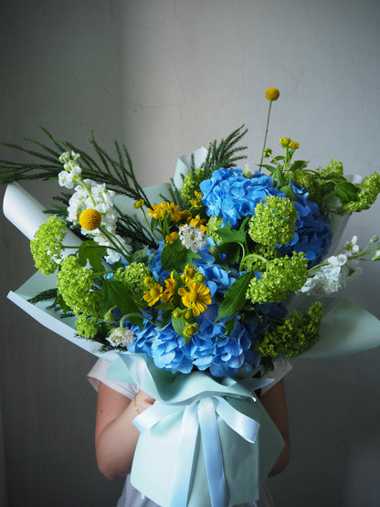 Bouquet of blue hydrangeas, greenery, and yellow flowers held against a neutral background