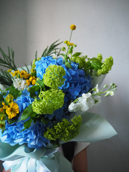 Bouquet of blue hydrangeas, green astilbes, and yellow daisies against a neutral background