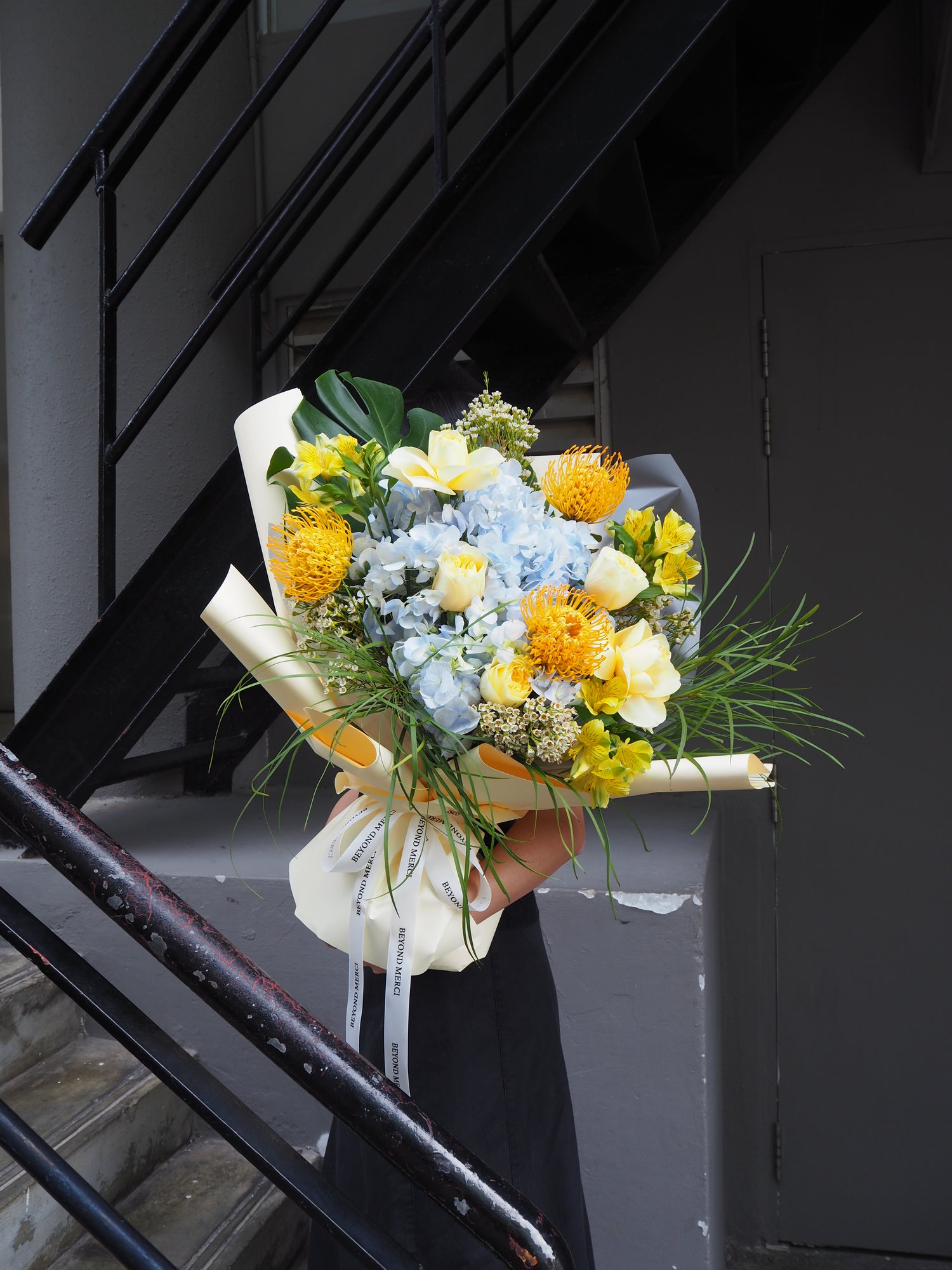 Grand fresh flower bouquet with a mix of pale blue hydrangea, yellow buttercup rose, pin cushion and green foliage.