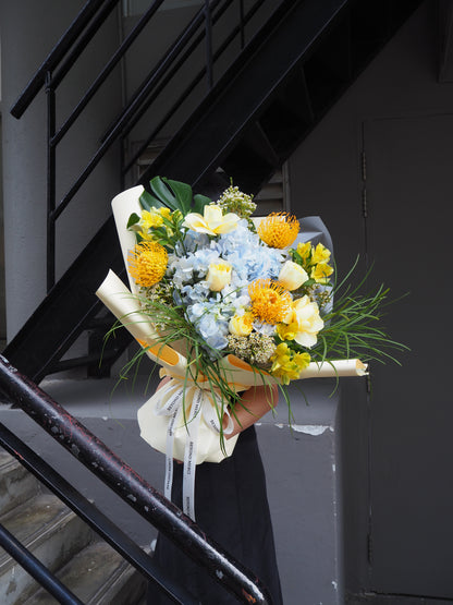 Grand fresh flower bouquet with a mix of pale blue hydrangea, yellow buttercup rose, pin cushion and green foliage.