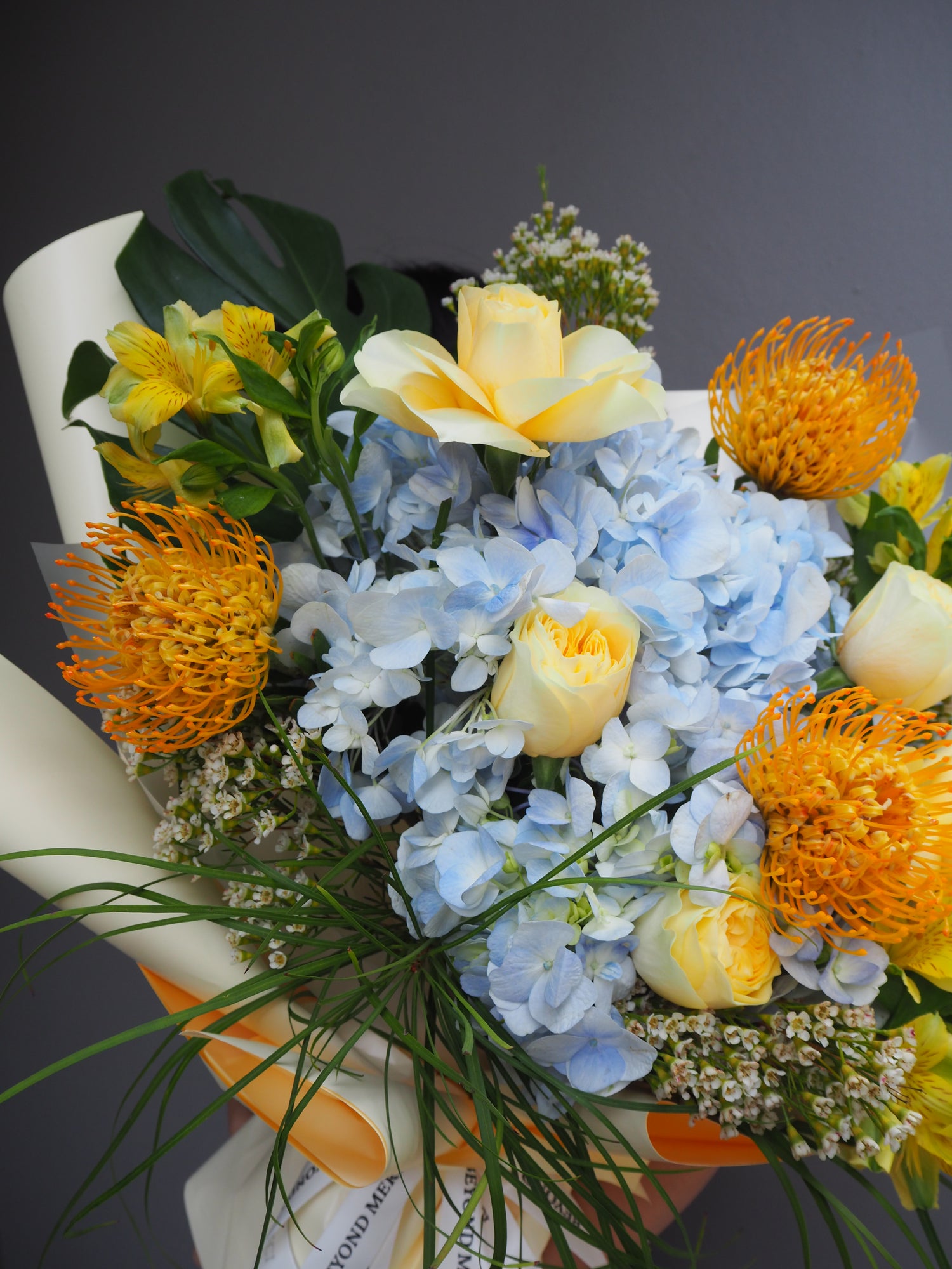 A luxe bouquet of flowers with blue hydrangeas, yellow roses, and yellow proteas on a dark background.