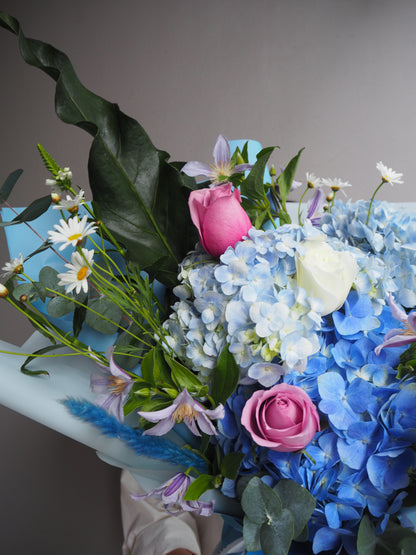 Large bouquet of blue hydrangeas, pink roses, and white daisies in a pale blue wrapper.