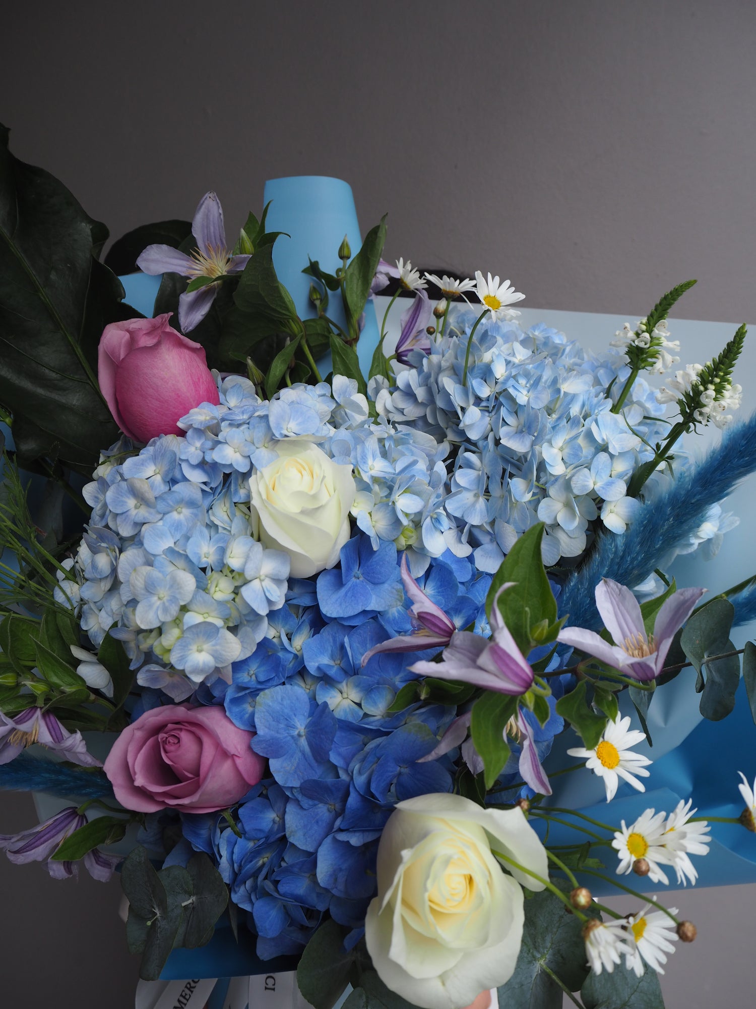 Fresh flower bouquet of flowers with blue hydrangeas, pink and white roses, and green leaves on a gray background.